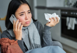 Portrait of ill young korean woman feeling sick, sneezing and holding napkin, staying at home ill, caught cold. Talking on mobile phone.