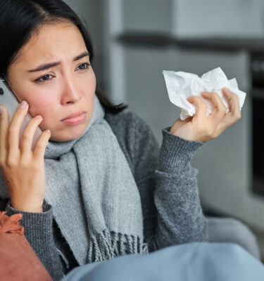 Portrait of ill young korean woman feeling sick, sneezing and holding napkin, staying at home ill, caught cold. Talking on mobile phone.