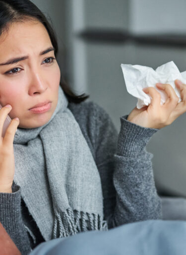 Portrait of ill young korean woman feeling sick, sneezing and holding napkin, staying at home ill, caught cold. Talking on mobile phone.