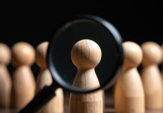 Magnifying glass examines wooden figure in a row of identical people.