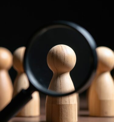 Magnifying glass examines wooden figure in a row of identical people.