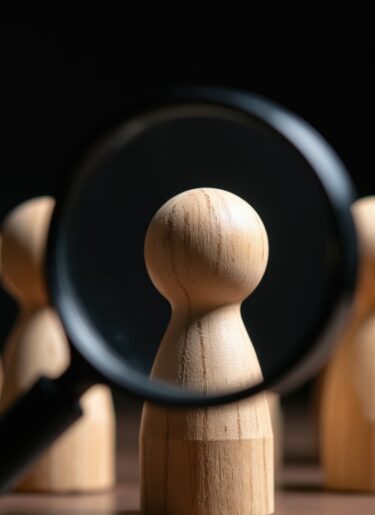 Magnifying glass examines wooden figure in a row of identical people.