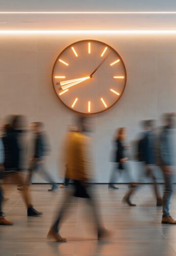 Blurry crowd of diverse people walking past a large illuminated clock in a modern office hallway during the workday