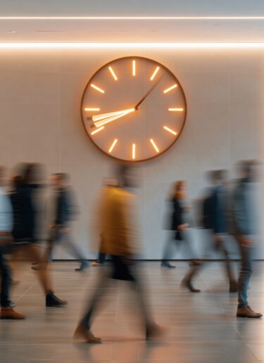 Blurry crowd of diverse people walking past a large illuminated clock in a modern office hallway during the workday