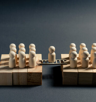 Wooden miniature figure standing on a bridge between two business teams, symbolizing mediation, negotiation, communication and organizational alignment.