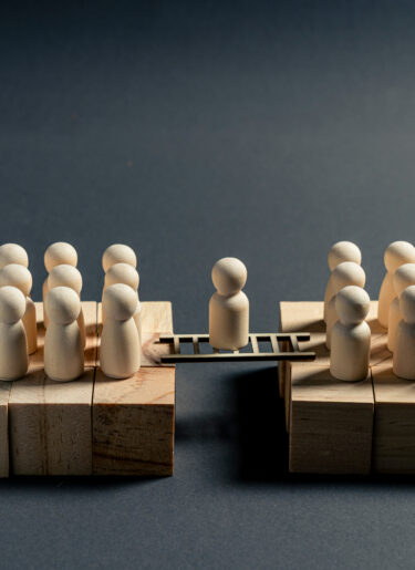 Wooden miniature figure standing on a bridge between two business teams, symbolizing mediation, negotiation, communication and organizational alignment.